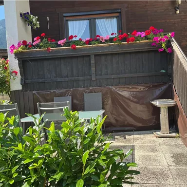 Eine gemütliche Terrasse mit einer grünen Hecke und einem Tisch mit Stühlen. Bunte Blumen schmücken den Balkon darüber.