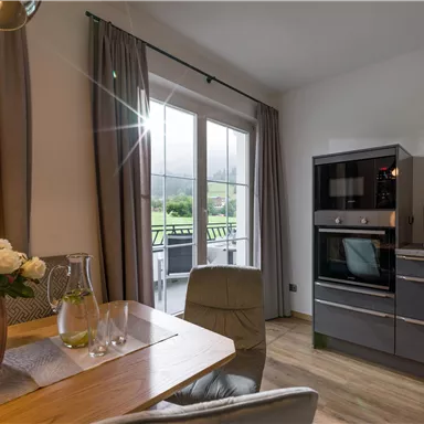 A modern kitchen with a dining area and large windows. There is a view of the outdoors and an attractive decoration with flowers.
