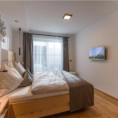 A modern bedroom with a large bed and wooden furniture. In front of the window, there are curtains and a television on the wall.
