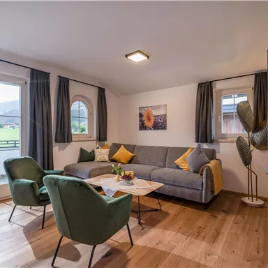 A modern living room with a gray sofa and green armchairs. Large windows offer a view of nature.