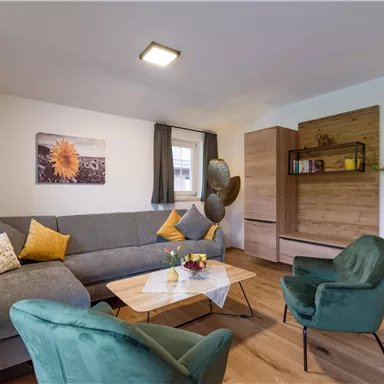 A modern living room with a gray sofa and green chairs. A table with flowers and a wooden shelf create a cozy atmosphere.