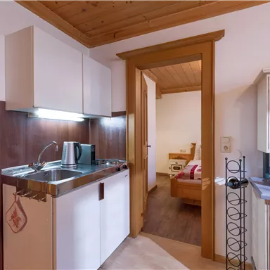Eine kleine, helle Küche mit modernen Möbeln und Holzdecke. Der Blick führt zu einem angrenzenden Raum mit einem gemütlichen Bett.
