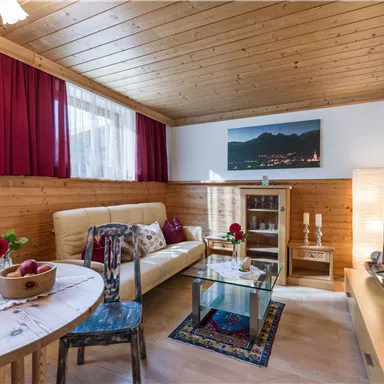 Ein gemütlicher Wohnraum mit Holzverkleidung, einem komfortablen Sofa und einem Tisch. Die Fenster lassen viel Licht herein, und es gibt dekorative Elemente wie Blumen und Kunstwerke.
