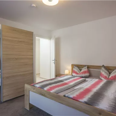 Ein gemütliches Schlafzimmer mit einem Doppelbett und stilvollen Kissen. Es gibt einen Holzschrank und viel natürliches Licht durch das Fenster.