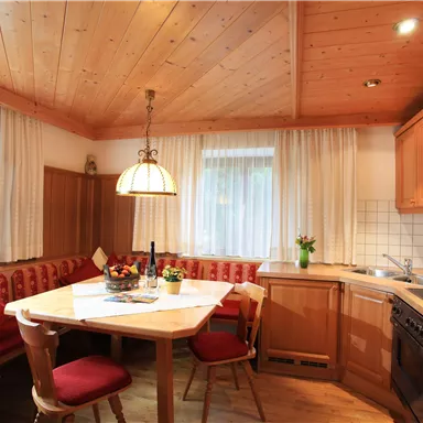 A cozy kitchen with wooden ceilings and a round table. Red upholstered chairs and a cooking area create an inviting atmosphere.