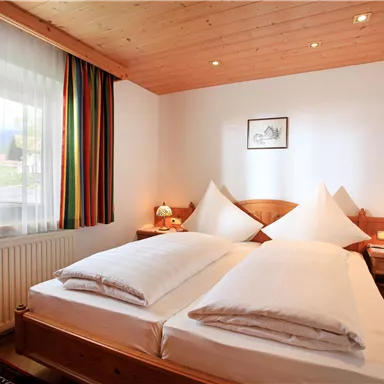 A cozy bedroom with two single beds and wooden decor. The room has large windows with curtains and two nightstands with lamps.