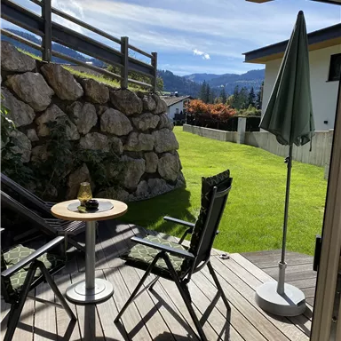 A beautiful balcony with two chairs and a table. In the background, there is a green garden and a cloudy mountain landscape.