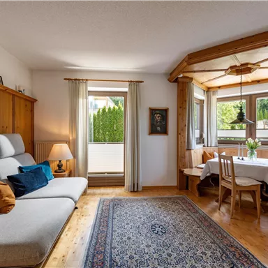 A cozy living room with a bright sofa and wooden shelves. The dining area has large windows with a view of the green surroundings.