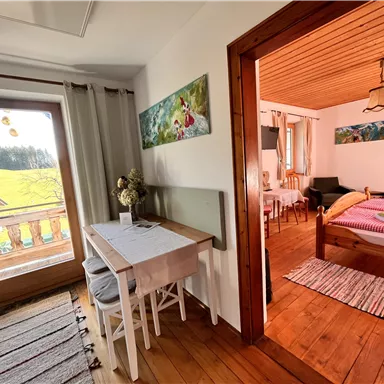 Ein gemütliches Zimmer mit Holzfußboden und einem Tisch. Durch das Fenster blickt man auf die grüne Landschaft.
