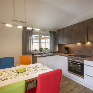 A modern kitchen with bright cabinets and a small dining table. Colorful chairs and windows create an inviting atmosphere.