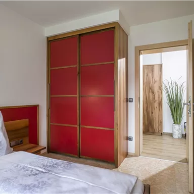 A cozy bedroom with a simple bed and a red wardrobe. In the background, a bright entrance area with a plant is visible.