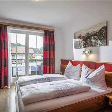 A cozy bedroom with a double bed and modern furniture. Large windows with red curtains offer a view outside.