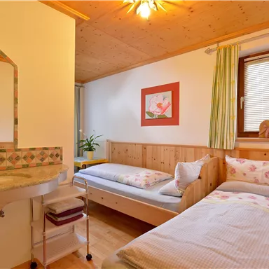 Ein gemütliches Zimmer mit zwei Einzelbetten und Holzverkleidung. Ein kleiner Waschbereich und ein Fenster sorgen für Helligkeit und eine einladende Atmosphäre.