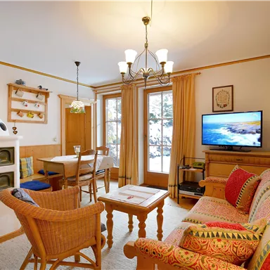 Ein gemütliches Wohnzimmer mit einem Sofa, einem Tisch und einem Fernseher. Die Raumgestaltung ist warm und einladend, mit einem Holzofen und viel natürlichem Licht.