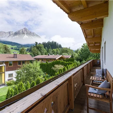 A cozy balcony with a wooden railing, overlooking green meadows and a majestic mountain in the background. The atmosphere is calm and inviting.