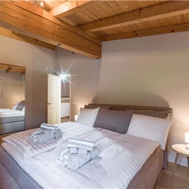 A cozy bedroom with wooden ceilings and a large bed. Soft lighting and a mirror create an inviting atmosphere.