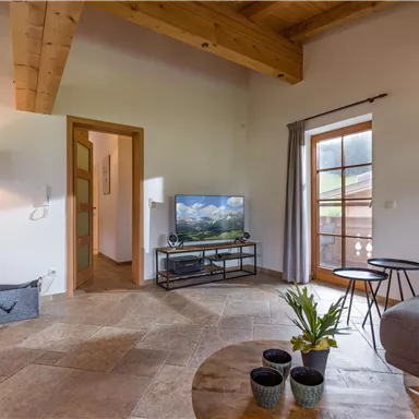A cozy living room with wooden beams and large windows. There is a TV, a sofa, and plants on a table.