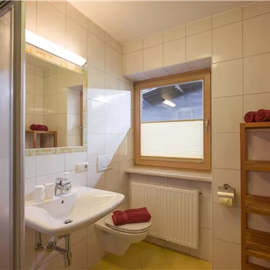 A bright bathroom with white tiles and a window. It contains a sink, a toilet, and a wooden shelf with towels.