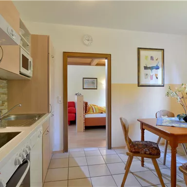 A bright kitchen with wooden cabinets and a table. In the background, a living room with a red sofa can be seen.