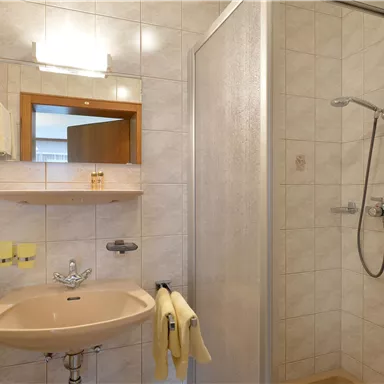 Ein helles Badezimmer mit einem Waschbecken, einem Spiegel und einer Dusche. Die Wände sind mit hellen Fliesen verkleidet.