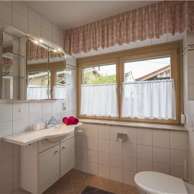 A bright bathroom with white tiles and a large window. Red towels hang on the wall, and a sink with a mirror is clearly visible.