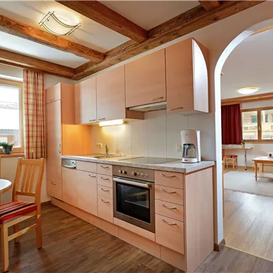 A bright kitchen with wooden shelves and modern appliances. In the background, there is a cozy living area with colorful curtains.
