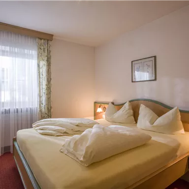A cozy bedroom with a large bed and two pillows. The windows are adorned with curtains and provide daylight.