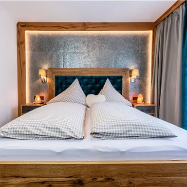 Ein gemütliches Schlafzimmer mit einem großen Bett und Holzrahmen. Die sanfte Beleuchtung und die hellen Vorhänge schaffen eine einladende Atmosphäre.