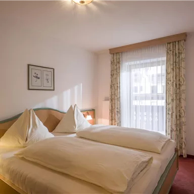 A cozy bedroom with two single beds and elegant bedding. The room is bright and inviting, with windows and curtains that let in natural light.