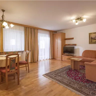 A cozy living room with a table, chairs, and a couch. Large windows let in plenty of daylight, and the floor is made of wood.