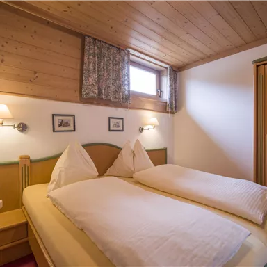 A cozy bedroom with a double bed and wooden walls. There are windows with curtains and a wardrobe.