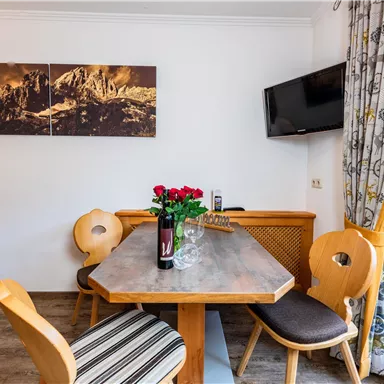 Ein gemütlicher Essbereich mit einem Holztisch und Stühlen. Auf dem Tisch stehen Rosen und eine Flasche, und im Hintergrund ist ein Fernseher und ein Gemälde zu sehen.