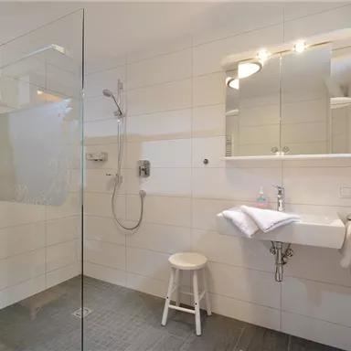 Ein modernes Badezimmer mit einer gläsernen Dusche und hellen Fliesen. Eine schöne Holzablage mit Handtüchern und ein kleiner Hocker vervollständigen den Raum.