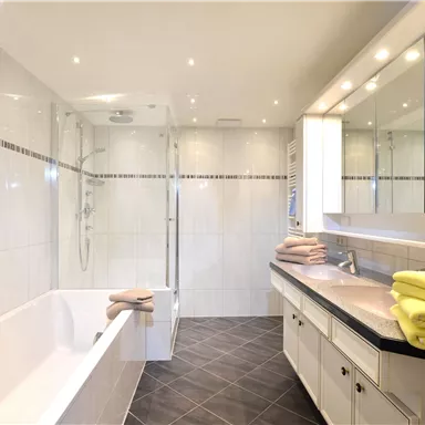 Ein modernes Badezimmer mit einer Badewanne und einer Dusche. Die Wände sind hell gefliest, und frische Handtücher liegen auf der Ablage.