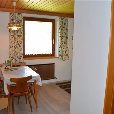 A cozy kitchen with a dining table and chairs. The walls are decorated with floral patterns and a window lets in plenty of light.