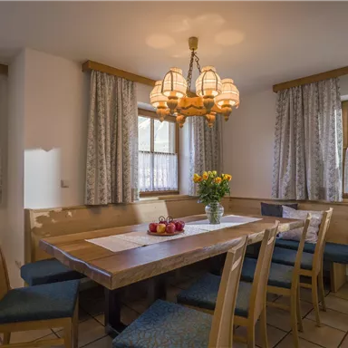 Ein gemütlicher Essbereich mit einem großen Holztisch und hellen Stühlen. Die Fenster lassen viel Tageslicht herein und eine Blumenvase verleiht dem Raum eine frische Note.