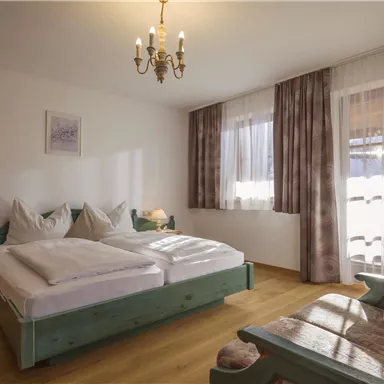 Ein gemütliches Schlafzimmer mit zwei Betten und einem Holzschränkchen. Große Fenster lassen viel Tageslicht herein.