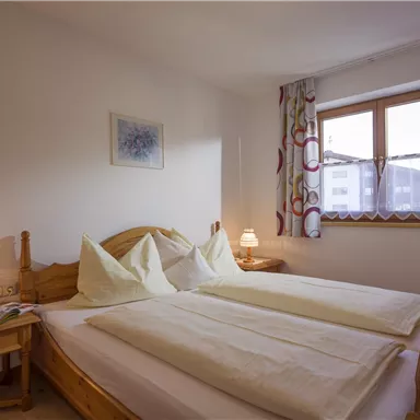 Ein gemütliches Schlafzimmer mit zwei Einzelbetten und sanften Farbtönen. Fenster mit Vorhängen lassen Tageslicht herein, und eine Nachttischlampe sorgt für eine angenehme Atmosphäre.