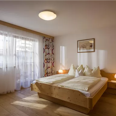 Ein gemütliches Schlafzimmer mit einem großen Bett und Holzdetails. Große Fenster sorgen für viel Tageslicht.