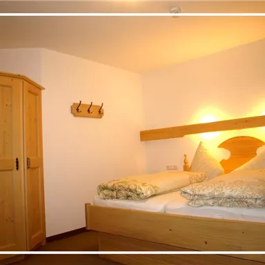A cozy bedroom with a double bed and decorative cushions. In the background, there is a wooden closet and a wardrobe.