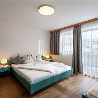 Ein modernes Schlafzimmer mit einem großen Bett und einem zusätzlichen Schlafplatz. Helle Vorhänge und Holzfußboden sorgen für eine angenehme Atmosphäre.