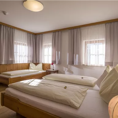 Ein helles Zimmer mit zwei Einzelbetten und Holzdetails. Die Fenster sind mit Vorhängen abgedeckt und es gibt eine gemütliche Stuhl.