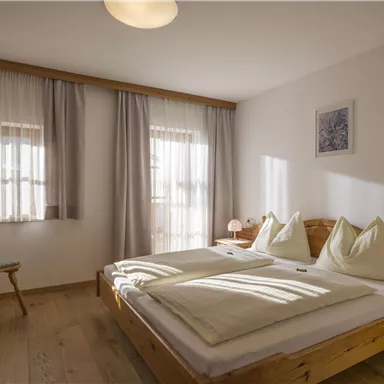 Ein helles Schlafzimmer mit einem Doppelbett und großen Fenstern. Die Einrichtung ist schlicht und gemütlich, mit einem Holzstuhl und sanften Vorhängen.
