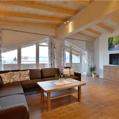 Ein modernes Wohnzimmer mit großen Fenstern und Holzböden. Es gibt ein gemütliches Sofa, einen Tisch und einen Fernseher.