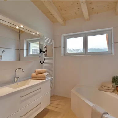 Ein modernes Badezimmer mit einer Badewanne und einem Waschbecken. Große Fenster sorgen für viel Tageslicht und frische Atmosphäre.