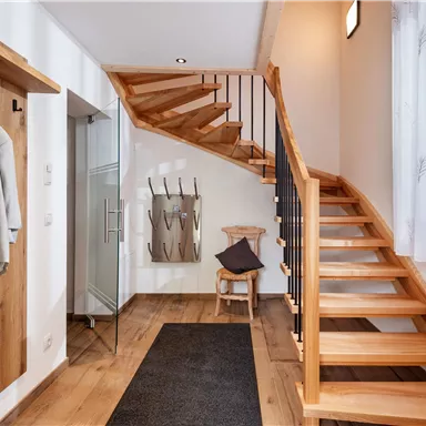 Ein einladender Eingangsbereich mit einer Holztreppe und hellem Licht. An der Wand hängen Jacken und eine Garderobe ist vorhanden.