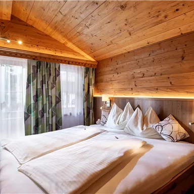 Ein gemütliches Schlafzimmer mit Holzdecke und warmem Licht. Das Bett ist einladend, umgeben von Fenstern und dekorativen Vorhängen.