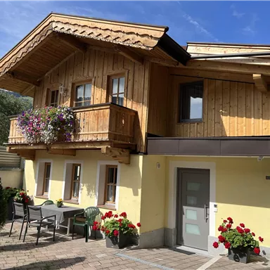 Ein gemütliches Holzhaus mit einem großen Balkon und bunten Blumen. Vor dem Haus steht ein Tisch mit Stühlen in einem schönen Garten.