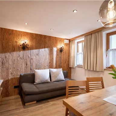 Ein gemütlicher Wohnraum mit einem grauen Sofa und einem Esstisch. Die Wände sind aus Holz, und es gibt große Fenster mit hellen Vorhängen.