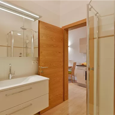 Ein modernes Badezimmer mit einem Waschbecken, Spiegeln und einer Dusche. Die Wände sind hell und es gibt eine Holztür, die zu einem weiteren Raum führt.